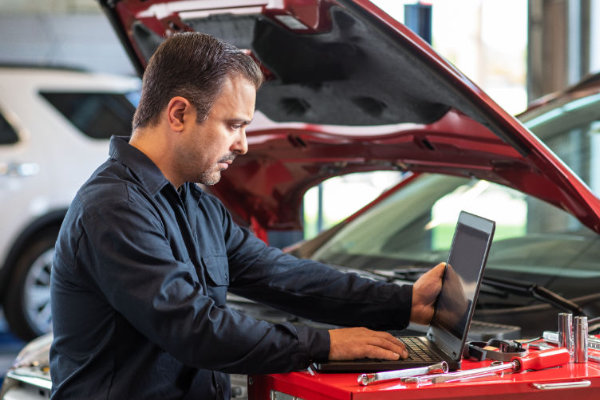 Automechaniker mit Laptop bei geöffneter Motorhaube eines roten Fahrzeuges