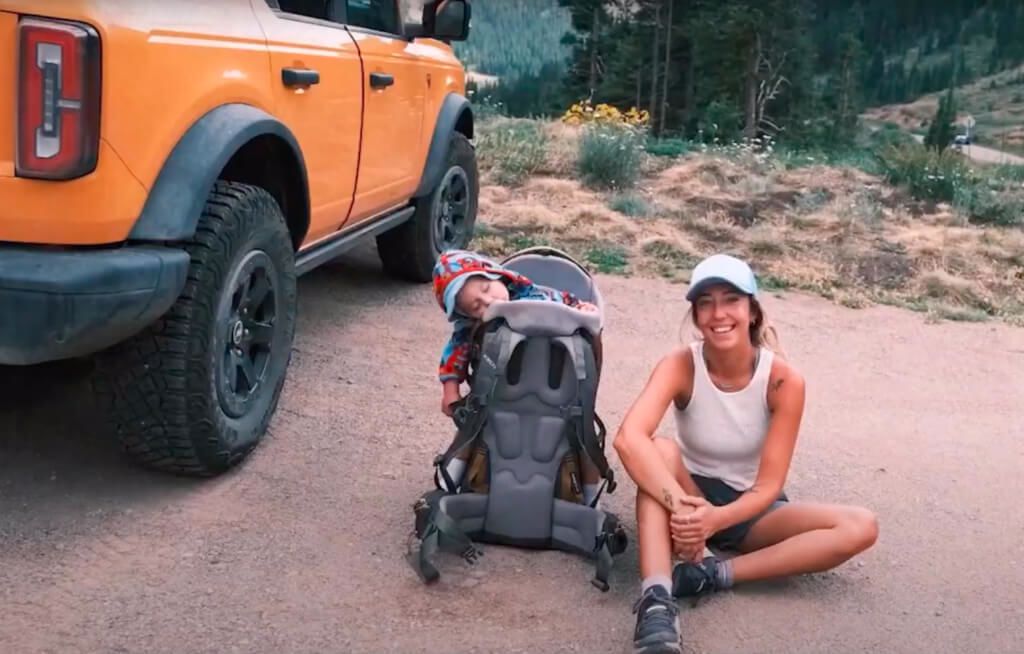 Mulher sentada no chão sorrindo com bebê ao lado e um Ford Bronco estacionado em ambiente off-road